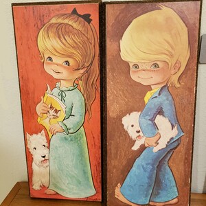 May include: Two framed illustrations of children with their pets. The girl on the left is wearing a blue nightgown and holding a book with a cat on the cover. She has a white dog by her side. The boy on the right is wearing blue pants and a blue shirt. He is holding a white dog in his arms. Both illustrations have a brown background and are signed "Jandros".