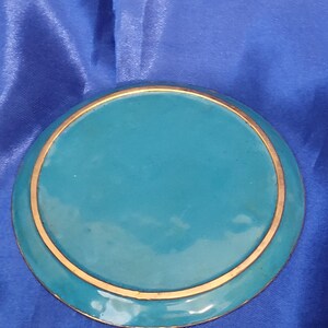May include: A round, teal-colored decorative plate with a gold-colored rim. The plate has a glossy finish and is set against a blue background. The design is simple and elegant.