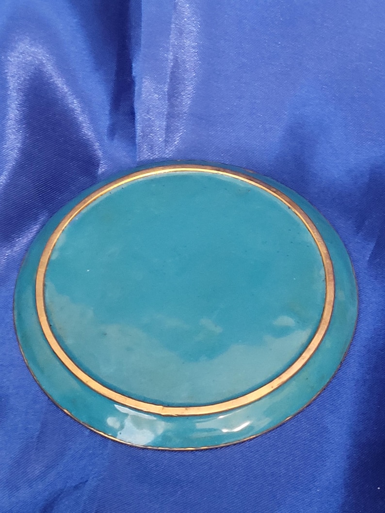 May include: A round, teal-colored decorative plate with a gold-colored rim. The plate has a glossy finish and is set against a blue background. The design is simple and elegant.