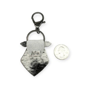 May include: A black and white cowhide leather keychain with a silver metal ring and clasp. The keychain is shaped like a cow's head and has a small, white leather flap on the top.