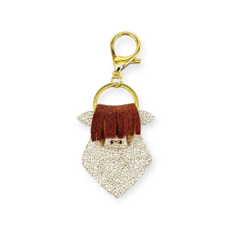 May include: A gold-colored keychain featuring a Highland cow design. The cow has a white, textured body, brown fringe hair, and small ears. The keychain includes a gold ring and clasp for easy attachment.