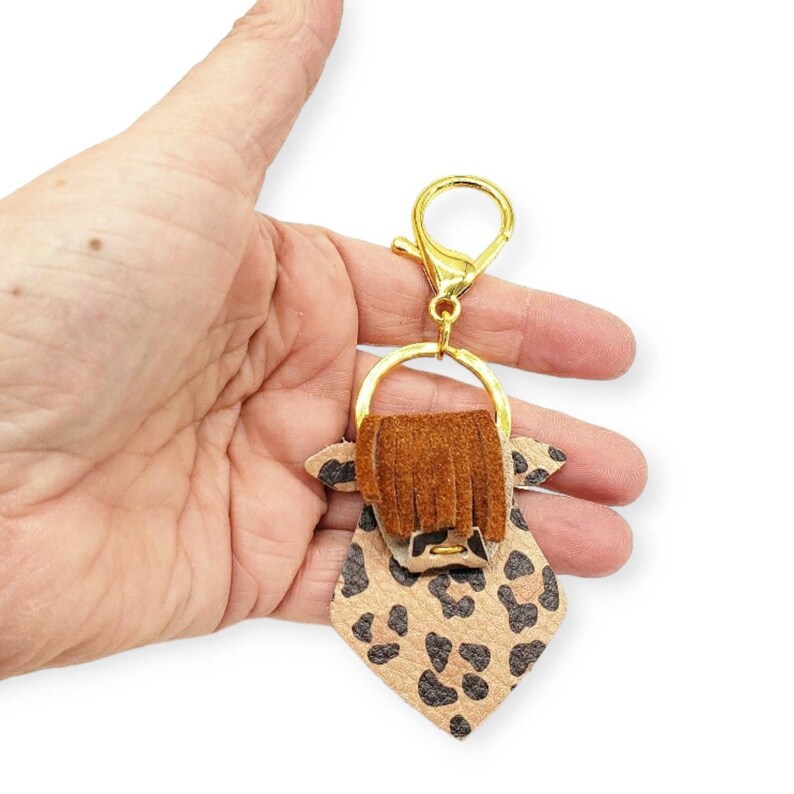 May include: A keychain featuring a cow design with a gold-colored clasp and ring. The cow has a tan and black leopard print body, brown fringe, and tan ears. The keychain is held in a hand against a white background.