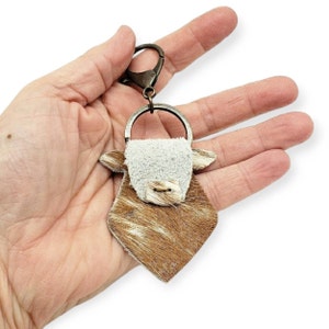 May include: A brown and white cowhide keychain with a white face and a silver ring. The keychain is attached to a silver key ring.
