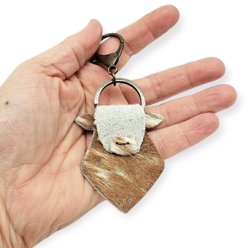 May include: A brown and white cowhide keychain with a white face and a silver ring. The keychain is attached to a silver key ring.