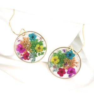 May include: A pair of round earrings with a gold-colored wire design. Each earring features a clear, circular resin pendant filled with colorful dried flowers in shades of pink, blue, yellow, and orange. The earrings are displayed on a white background.