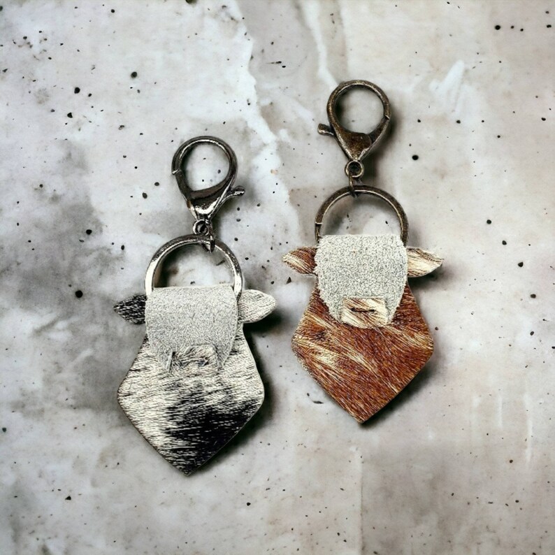 May include: Two cow-shaped keychains with silver key rings. One keychain is brown and white, the other is silver and white.