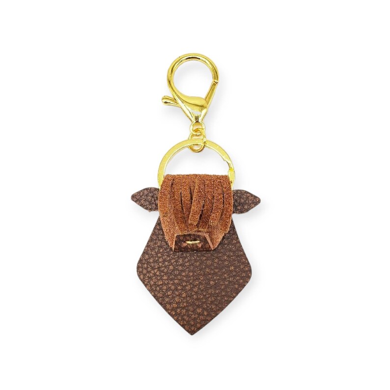 May include: A brown leather keychain in the shape of a Highland cow. The keychain has a gold-colored clasp and ring. The cow has a textured body, fringed bangs, and small ears. A unique accessory for keys or bags.