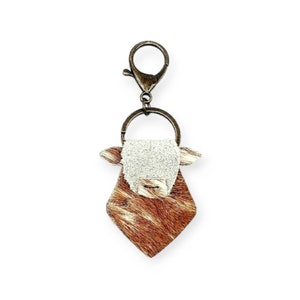May include: A brown and white cowhide keychain with a metal ring and clip. The cowhide is cut in a diamond shape with a white cow face in the center.