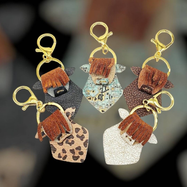 May include: Keychains in the shape of Highland cows, each with a gold-colored clasp and ring. The cow keychains feature various textures and colors, including black, turquoise, brown, leopard print, and white, with brown fringe details.