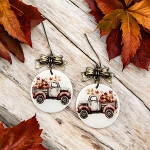 May include: A pair of round earrings featuring a white background with a vintage truck design carrying pumpkins and fall foliage. The earrings have a bronze bow and a wire hook. Autumn leaves are in the background.