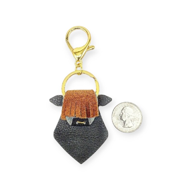 May include: A black and brown leather keychain in the shape of a Highland cow. The keychain has a gold-colored clasp and ring. The cow has a brown fringe and small gray ears. A quarter is next to the keychain for size comparison.