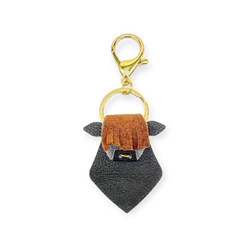 May include: A black leather keychain in the shape of a Highland cow, featuring a gold-colored clasp and ring. The cow has brown fringe, gray horns, and a diamond-shaped body. A unique accessory.