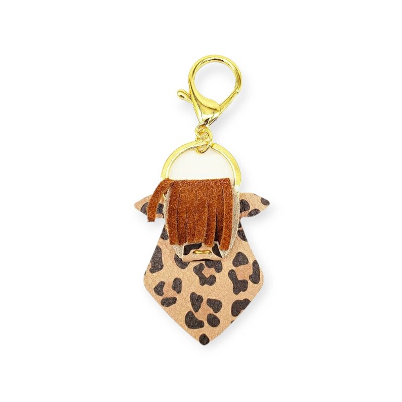 May include: A keychain featuring a tan and black leopard print design with a brown fringe and gold-colored hardware. The keychain has a gold-colored clasp and a ring for attaching keys.