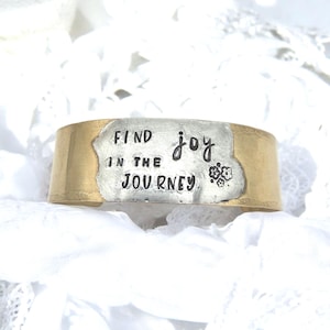 May include: A wide, gold-toned metal cuff bracelet with a silver-toned center. The center features the words "FIND joy IN THE JOURNEY" in black lettering, along with a small floral design. The bracelet is set against a white, textured background.