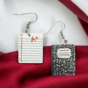 May include: A pair of silver earrings with a notebook and a sheet of paper charm. The notebook charm has the word "COMPOSITION" printed on it. The sheet of paper charm has an "A+" written in red.