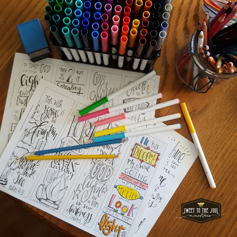 May include: A set of printable bookmarks with inspirational quotes and bible verses. The bookmarks are black and white with a hand-lettered style. The image shows a variety of colored markers and pencils for coloring the bookmarks.