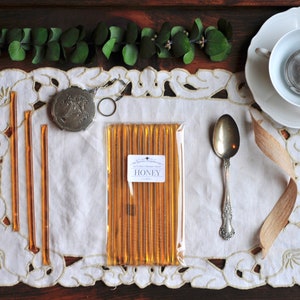 May include: A white doily with a gold trim sits on a wooden surface.  A package of honey sticks is in the center of the doily.  The package is labeled "HONEY".  There is a silver spoon, a small silver compact, and a teacup and saucer on the doily.
