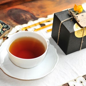 May include: A white teacup and saucer with a cup of dark tea. A black box with a brown twine bow and a tag that reads "Tea Sampler Loose Tea" is on a white tablecloth with a lace trim.