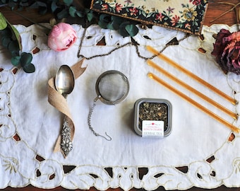 May include: A silver tea infuser with a chain, a silver spoon wrapped in burlap ribbon, a small tin of "Authentic Kings Charm Herbal Tea", and three honey sticks on a white lace tablecloth with pink and dried flowers.