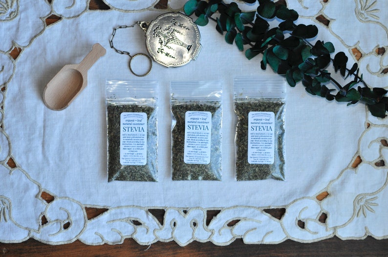 May include: Three small clear plastic bags filled with dried green stevia leaves. The bags are labeled "organic leaf natural sweetener stevia". The bags are on a white lace tablecloth with a wooden spoon and a silver tea strainer.