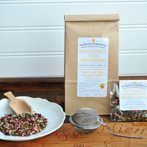 May include: A brown paper bag with a label that reads "The Queen's Tea Apothecary, Queen's Traditions, Tulsi Ashwagandha, Loose Herbal Infusion - Caffeine Free, Organic Ashwagandha, Organic Tulsi, Organic Green Tea, Organic Rose, Organic Ginger, Organic Eleuthero, Organic Dried Orange Peel" and a small clear plastic bag with a label that reads "The Queen's Tea Apothecary, Queen's Traditions, Tulsi Ashwagandha, Loose Herbal Infusion - Caffeine Free"  A white plate with a wooden spoon and a loose leaf tea blend. A silver tea infuser.