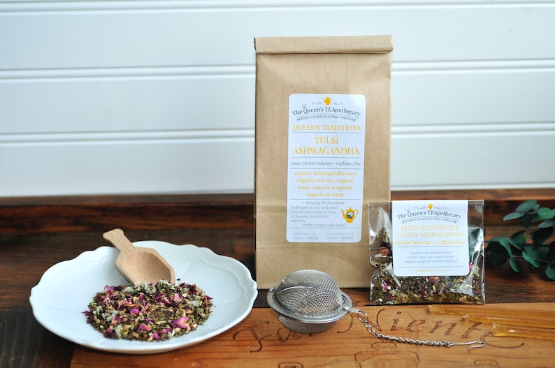 May include: A brown paper bag with a label that reads "The Queen's Tea Apothecary, Queen's Traditions, Tulsi Ashwagandha, Loose Herbal Infusion - Caffeine Free, Organic Ashwagandha, Organic Tulsi, Organic Green Tea, Organic Rose, Organic Ginger, Organic Eleuthero, Organic Dried Orange Peel" and a small clear plastic bag with a label that reads "The Queen's Tea Apothecary, Queen's Traditions, Tulsi Ashwagandha, Loose Herbal Infusion - Caffeine Free"  A white plate with a wooden spoon and a loose leaf tea blend. A silver tea infuser.