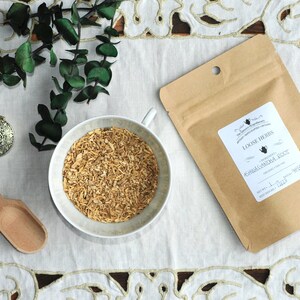 Op de afbeelding: Een bruine papieren zak met een wit label waarop staat "Loose Herbs, Ashwagandha Root, Net Wt. 1 oz (28g)". De zak ligt op een wit kantkleed met een kleine witte kom gevuld met gedroogde kruiden en een houten lepel.