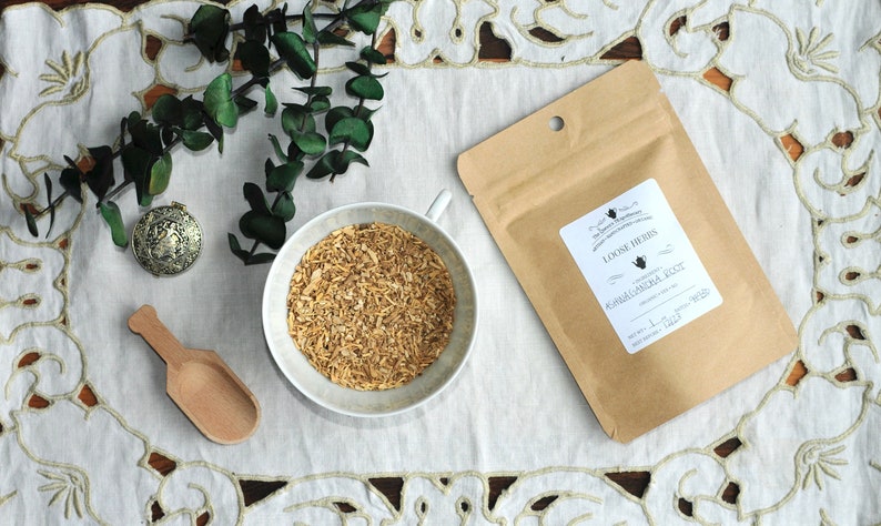 Op de afbeelding: Een bruine papieren zak met een wit label waarop staat "Loose Herbs, Ashwagandha Root, Net Wt. 1 oz (28g)". De zak ligt op een wit kantkleed met een kleine witte kom gevuld met gedroogde kruiden en een houten lepel.