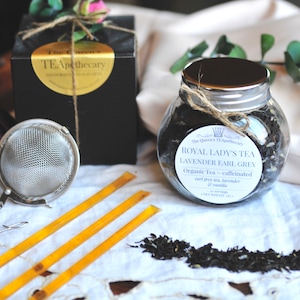 May include: A glass jar filled with loose leaf tea, labeled "Royal Lady's Tea Lavender Earl Grey Organic Tea - caffeinated earl grey tea, lavender & vanilla". The jar is sitting on a white lace tablecloth with three yellow sticks and a silver tea strainer.