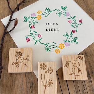 Może przedstawiać: Kwiatowa kartka z pozdrowieniami z napisem "Alles Liebe" otoczonym wieńcem z zielonych liści, różowych i żółtych kwiatów. Obecne są również trzy drewniane stemple z kwiatowymi wzorami.