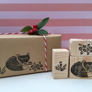 Op de afbeelding: Een set van drie houten stempels met een slapende vos, een bloem en een enkele stengel met bladeren. De stempels bevinden zich op een bruin papieren pakket met rood en wit touw.