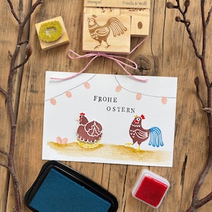Könnte beinhalten: Oster-Bastelzubehör mit Holzstempeln mit Hühnerdesigns, einer Grußkarte mit dem Text "FROHE OSTERN" und Stempelkissen. Die Stempel sind mit rosa Bindfaden zusammengebunden. Maße in cm.
