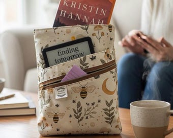 Op de afbeelding: Een crèmekleurige boekenhoes met een rits, met een patroon van bijen, paddenstoelen en bladeren. Een boek van Kristin Hannah en een e-reader met de tekst "finding tomorrow" zijn zichtbaar. Een klein paars notitieblokje zit in de voorzak.