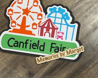 May include: A die-cut sticker featuring a Ferris wheel, roller coaster, and a fair tent. The words "Canfield Fair" are printed in black on a green background. The sticker also includes the text "Memories by Margot".