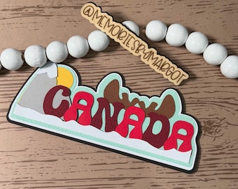 May include: A layered paper craft featuring the word "CANADA" in red, with a mountain and moose design. A wooden sign with "@MEMORIESBYMARGOT" is above the craft, and white beads are in the background. The craft is on a wooden surface.