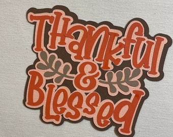 May include: A brown and orange paper craft with the words "Thankful & Blessed" in a decorative font. The words are outlined in brown and have a textured appearance.