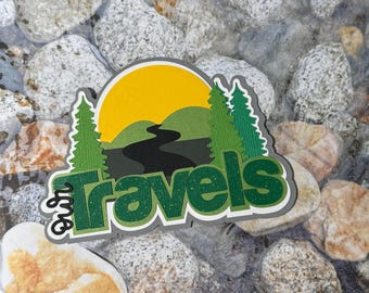 May include: A green and gray paper craft with the words "our travels" and a graphic of a sun, mountains, and a winding road.