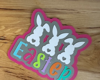 May include: A pink paper Easter craft with three white bunny silhouettes and the word "Easter" in colorful letters.