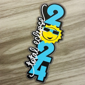 May include: A vertical cutout design with the text "total eclipse 2024". The numbers "2024" are in light blue, with a smiling sun wearing sunglasses in the center. The text "total eclipse" is in white cursive.