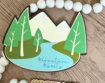 May include: A layered wooden mountain scene with green trees, a blue river, and white mountains. The phrase "Adventure Awaits" is written in blue script. The bottom of the image has the text "@MEMORIESBYMARGOT".