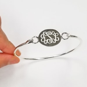 May include: A silver-toned bangle bracelet featuring a central oval-shaped charm with an intricate, cut-out monogram design. The bracelet has a thin, polished metal band that connects to the central charm with small circular jump rings.