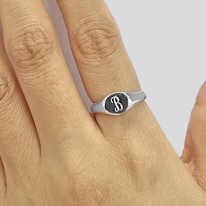 Könnte beinhalten: Ein silberner Siegelring mit einem schwarzen, ovalen Feld, das den Buchstaben "B" in Weiß zeigt. Der Ring wird an einem Finger getragen und unterstreicht sein schlichtes, aber elegantes Design. Ein klassisches Schmuckstück.