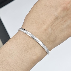 May include: A silver herringbone bracelet. The bracelet is a continuous band of interlocking, flat, rectangular links. The bracelet is worn on a wrist.