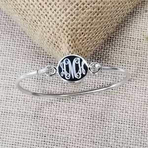 Op de afbeelding: Een zilveren armband met een ronde zwarte bedel met een monogram. De bedel is versierd met twee kleine heldere strass steentjes.