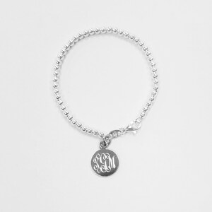Könnte beinhalten: Ein silbernes Armband mit Perlen und einem runden Anhänger mit einem Monogramm. Der Anhänger ist mit einem silbernen Verschluss am Armband befestigt.