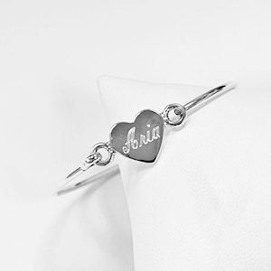 May include: A silver-toned bangle bracelet featuring a central heart-shaped charm. The heart charm is engraved with the name Aria in a cursive script. The charm is connected to the thin, polished metal band by two small circular jump rings.