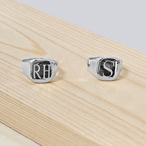 May include: Two silver signet rings with initials "RH" and "S" engraved on them. The rings are on a light brown wooden surface.