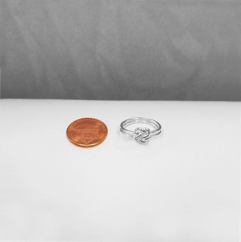 May include: A silver knot ring with a simple design. The ring is shown next to a US penny for size comparison.