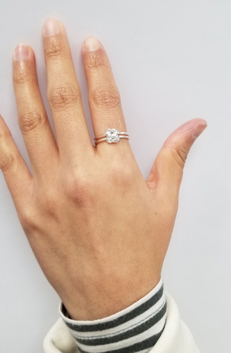 May include: A silver ring with a knot design. The ring is worn on the ring finger of a person's hand.