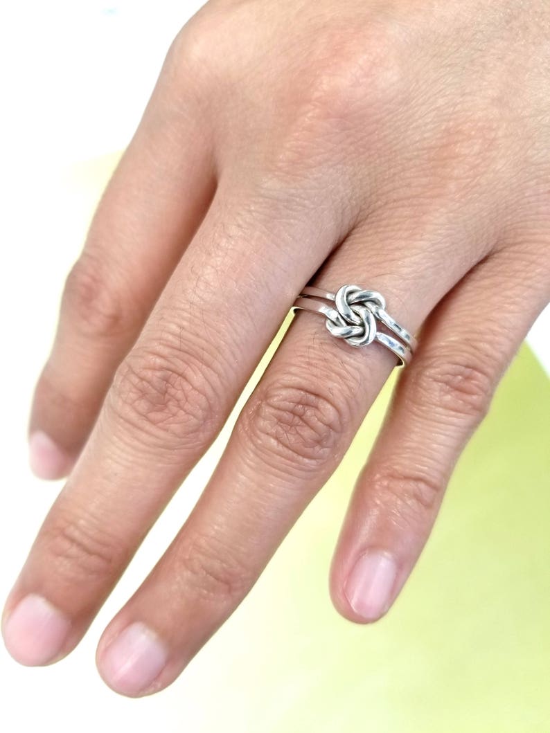 May include: A silver ring with a knot design. The ring is worn on the middle finger of a hand.